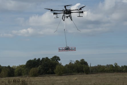 A latge drone flies in a field with a platfrom of equipment suspended beneath it.