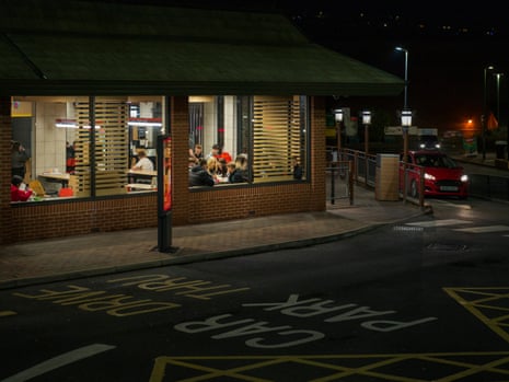 A McDonald’s restaurant seen from across the street at night. People can be seen inside through the lit-up windows