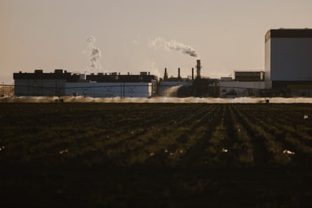 A farm field with an industrial facility spewing smoke in the background.