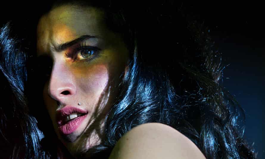 A close-up of Amy Winehouse looking over her shoulder at something off camera, her face tinted by coloured stage lights