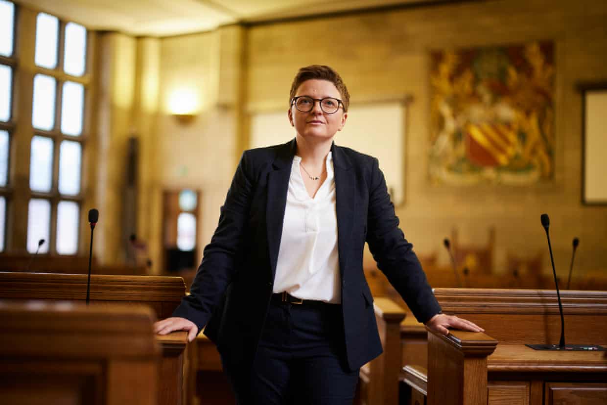 Bev Craig, Manchester council leader, stands between wooden benches in a council chamber