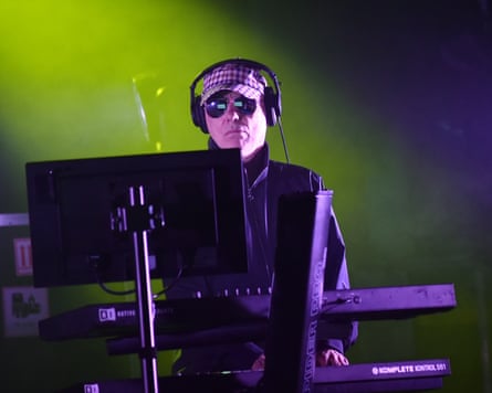 Chris Lowe wearing headphones, a hat and sunglasses and standing at a synth console