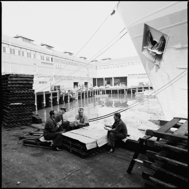 A black and white archival photograph depicts wharfies playing cards, Walsh Bay, c1970