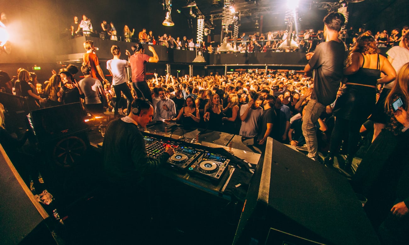 A crowd of dance music fans on the dancefloor at Amsterdam club De Marktkantine. The image is taken from behind the DJ booth, where a DJ is using digital decks to play music.