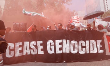 In a cloud of pink gas, a group of people hold a black banner that says in white letters Cease Genocide.