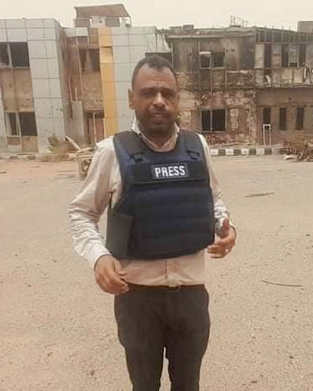 A man wearing a press vest stands in front of damaged buildings