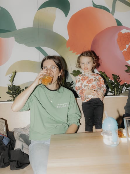 A seated woman in a mint sweater sips on a drink, a baby standing next to her