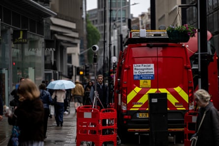 A red van with a sign saying “Live facial recognition in operation” is parked on a street in the City of London, with a tall camera on a tripod mounted on the pavement