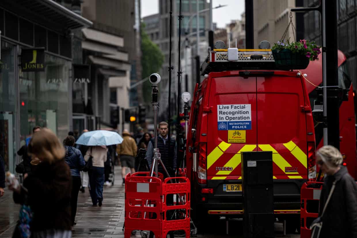 A red van with a sign saying “Live facial recognition in operation” is parked on a street in the City of London, with a tall camera on a tripod mounted on the pavement