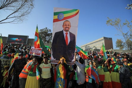 A crowd of women and men, many in Ethiopian flag colours, stand under a cloudless sky holding a vertical banner of Abiy