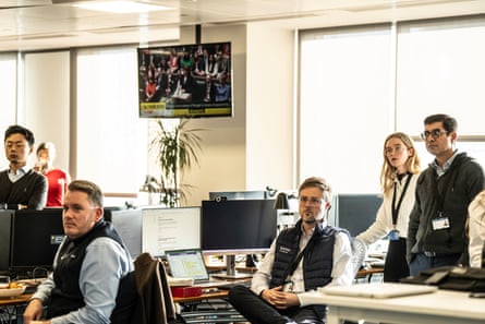 Traders sit in their office and stand by their desks, watching a screen. They are mostly quite young, male and female, and casually dressed; one man wears a gilet with Saxo logo. Another TV screen is on the wall behind them showing the budget announcement