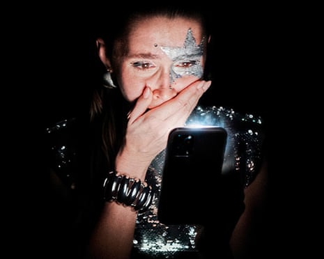 A woman with glittery face paint covers her mouth while looking at an illuminated phone screen in darkness, in The Influencer Project