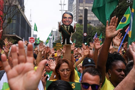 a crowd packs into a city centre street waving green and yellow Brazilian flags and holding up their hands; one person holds up a placard with an image of Bolsonaro smiling and holding up his hands in V for victory signs. The crowd appears to include young and older people, black and white.
