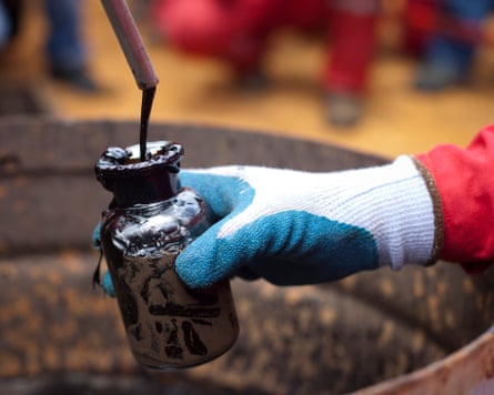 A gloved hand holding a jar to collect a sample of oil from a thin pipe