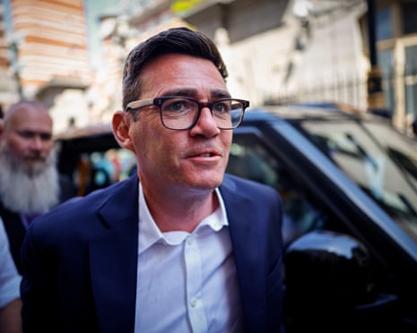 Andy Burnham in glasses and a dark suit looks upward in front of a blurred street scene