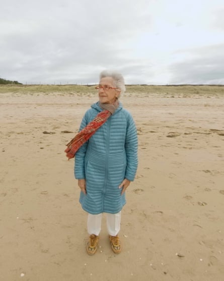 Marion Deichmann in long blue quilted anorak and white trousers with red scarf blowing in the wind, standing on a wintry beach
