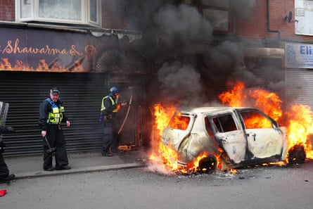 Flames and black smoke engulf a charred white Mini as police stand close by