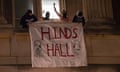 Nighttime photo of four young people with faces covered, holding a large white cloth sign out a building window that says "Hind's Hall"