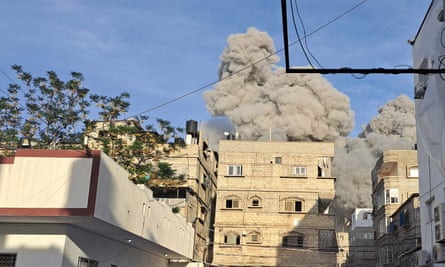 Smoke and dust billows in a huge cloud above buildings against blue daylight sky