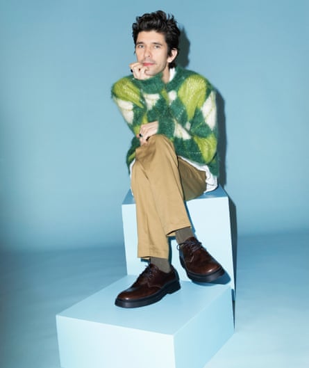 Actor Ben Whishaw in yellow and green jumper and brown trousers, sitting on white blocks, against blue background, January 2022