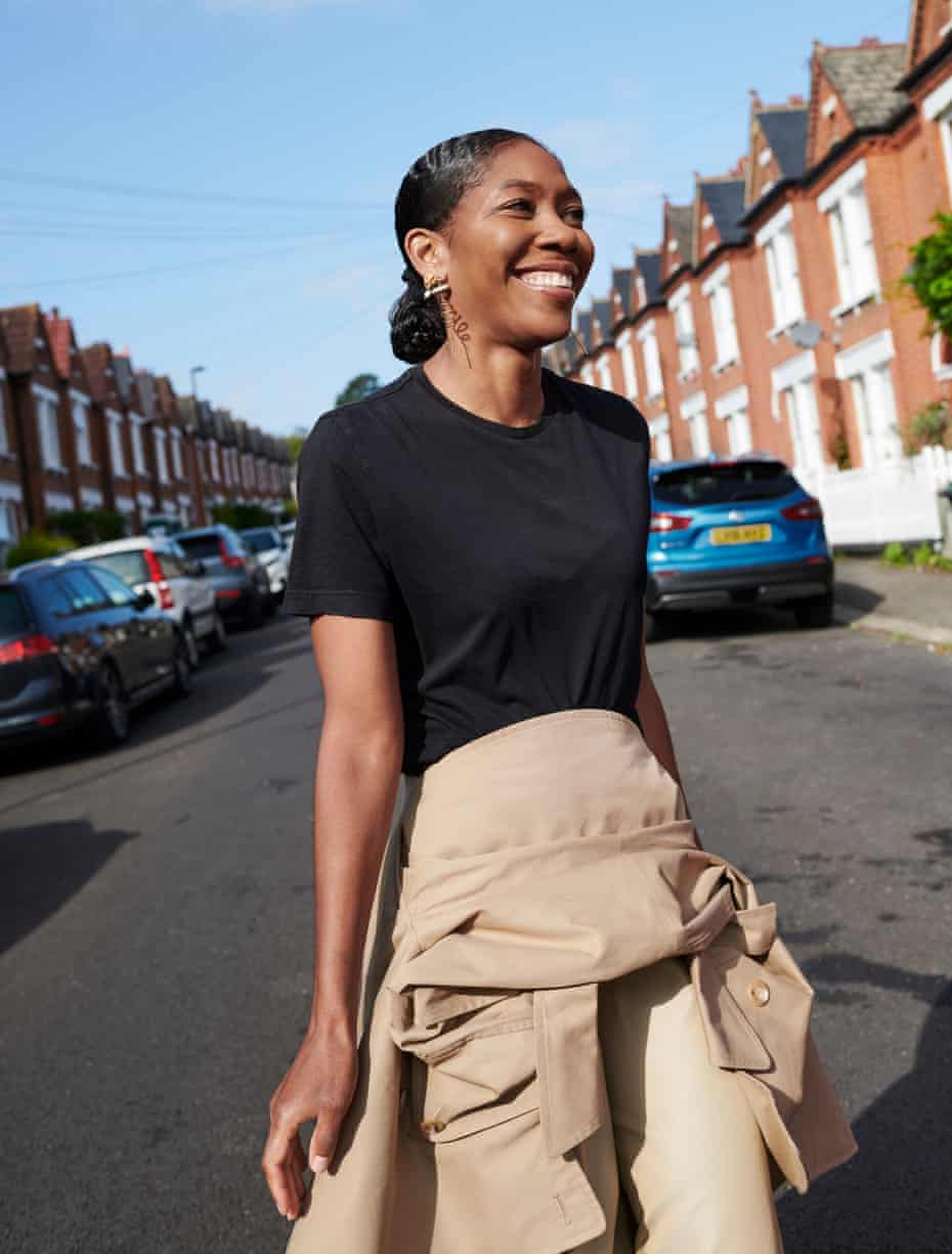 Kenya Hunt in a black T-shirt and with a coat tied around her waist, walking across a road with a row of houses behind her