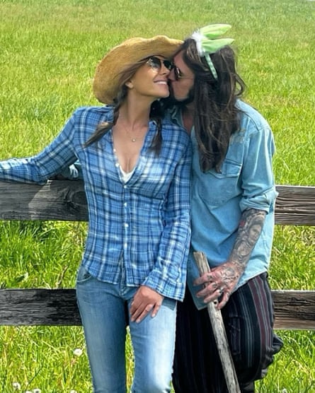 Elizabeth Hurley and Billy Ray Cyrus smooching and leaning against a fence with grass behind - she wearing a blue checked shirt, blue jeans and straw hat, he in a blue jeans shirt and striped trousers and fairy-style hairband