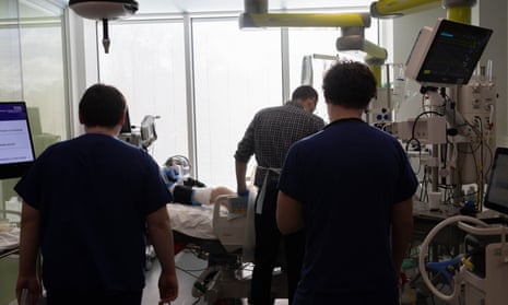 Three doctors silhouetted against a window looking at a patient in a hospital bed with equipment surrounding them.