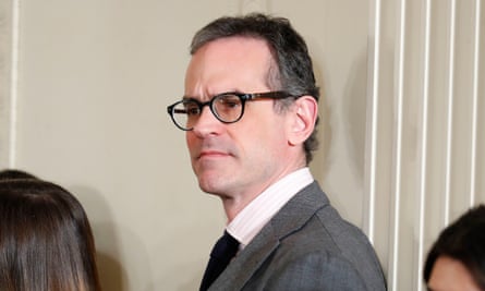 A middle-aged white man with dark gray hair combed back from his forehead and black-framed glasses, wearing a gray suit, stands against a white wall inside somewhere, looking serious.