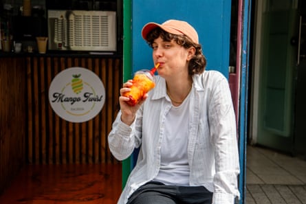 A woman wearing a pink cap and a white shirt and Tshirt, drinking a red and orange drink out of a plastic container