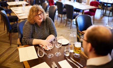 A young, balding man with glasses, wearing a grey top over a white shirt, and an older woman with curly red hair and glasses, wearing a grey sparkly top, sitting at a wooden restaurant table talking