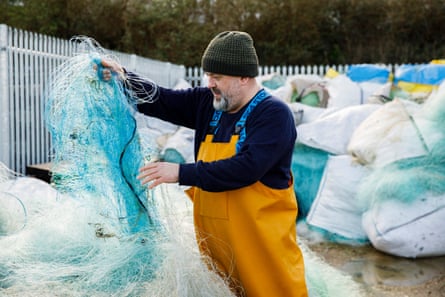A man in fishing overalls standing in a yard among large bags full of fishing nets