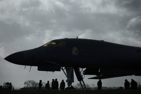 कुछ कर्मी एक बड़े बमवर्षक के नीचे खड़े हैं, हर कोई छाया में और अंधेरे में है क्योंकि वे भूरे आकाश के सामने छायांकित हैं।