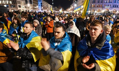 Big crowd at night, many draped in or carrying Ukraine flag
