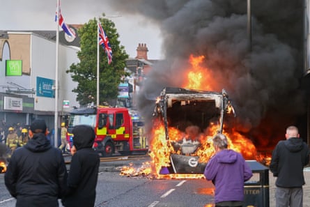People look on as a lorry burns in the middle of a road and firefighters arrive at the scene