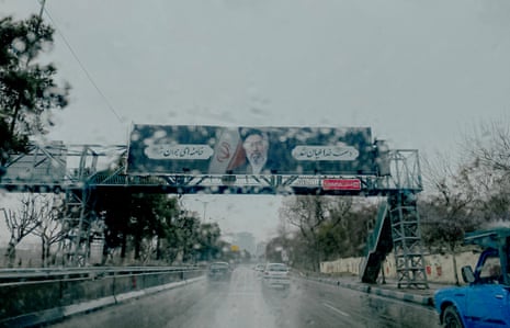 A rainy urban road in Tehran, with a gantry above it with images of religious leaders, pictured from inside a vehicle.