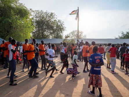 A crowd of people including dancing children gather on dusty ground next to a building and flagpole