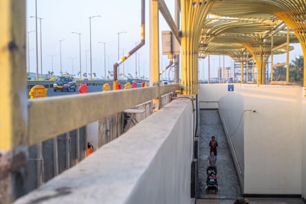 A woman pushes a child in a pram into a highway underpass under a yellow tubular metal sunshade