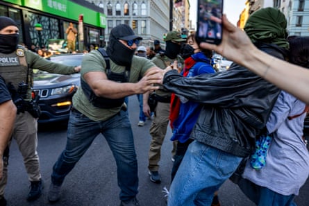 a masked man wearing a tactical vest reaches for a person on the street