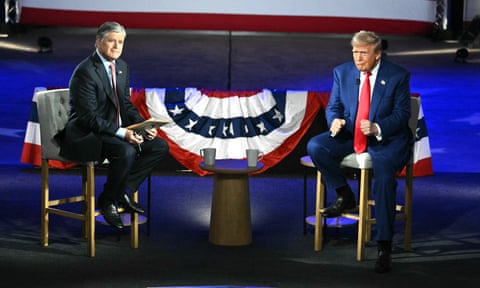 Two men sitting on a stage and the American flag can be seen between them