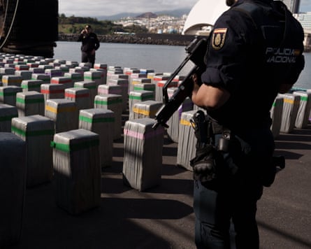 A police officer with a machine gun standing guard over rectangular bales wrapped in plastic at a quayside