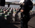 A Spanish policeman guards packages of cocaine on a quay, standing over them with a gun after they have been unloaded from a cargo ship.