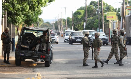 Security guards in combat gear holding rifles patrol a street with cars on