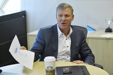 A man in a blue suit jacket with a white shirt open at the throat displays a piece of paper while sitting at a table. On the table in front of him is a Starbucks cup.