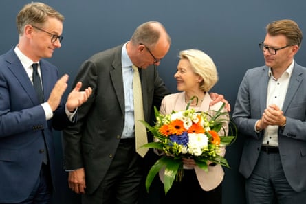 Friedrich Merz puts an arm around von der Leyen as she holds a bouquet of flowers. Two men in suits stand either side applauding