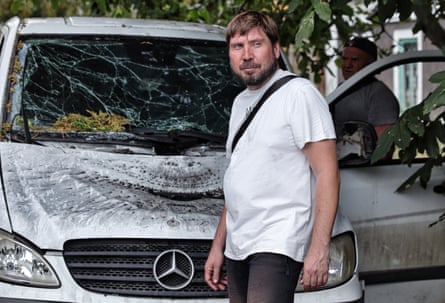 A man walks past a white Mercedes car with its windscreen shattered and bonnet dented and blackened.