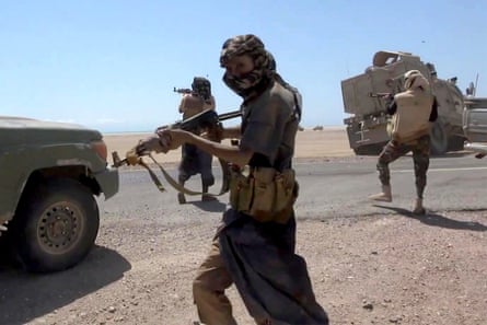 Fighters with machine guns walk across a road in a flat, sandy landscape with water in the background. They have scarves around their heads; one wears camouflage trousers and military-style boots. An armoured vehicle and pickup trucks are at the roadside.