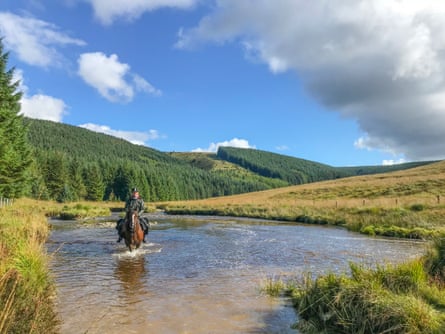 man riding horse in shallow river surrounded by green hills