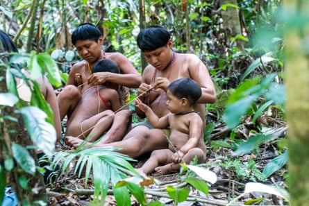 Two women sit with children in a clearing in dense forest, weaving.