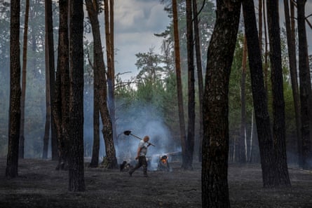A man walks through a charred forest with smoke behind him, a shovel over his shoulder