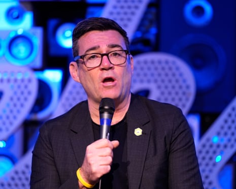 Andy Burnham speaks into a microphone against a blue-lit backdrop.